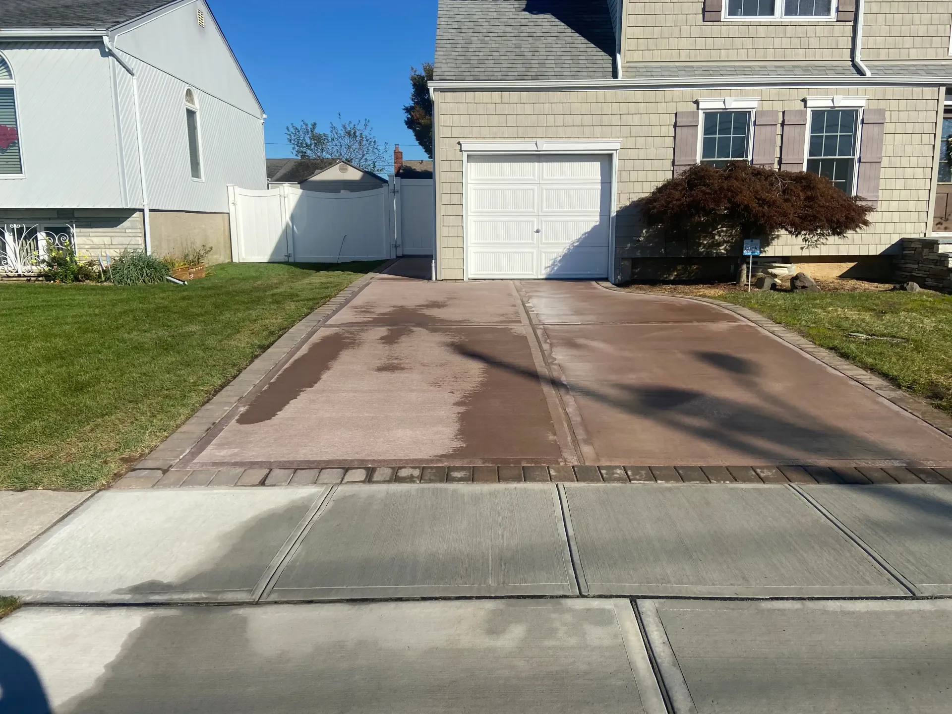 Driveway in front of a house with a garage; sidewalk in foreground. Brick edging, brown and tan surfaces.