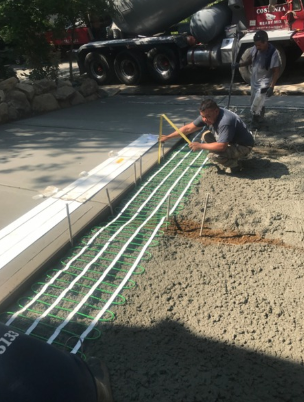 Workers install heating cables in wet concrete, using a tape measure outdoors. A concrete truck is in the background.
