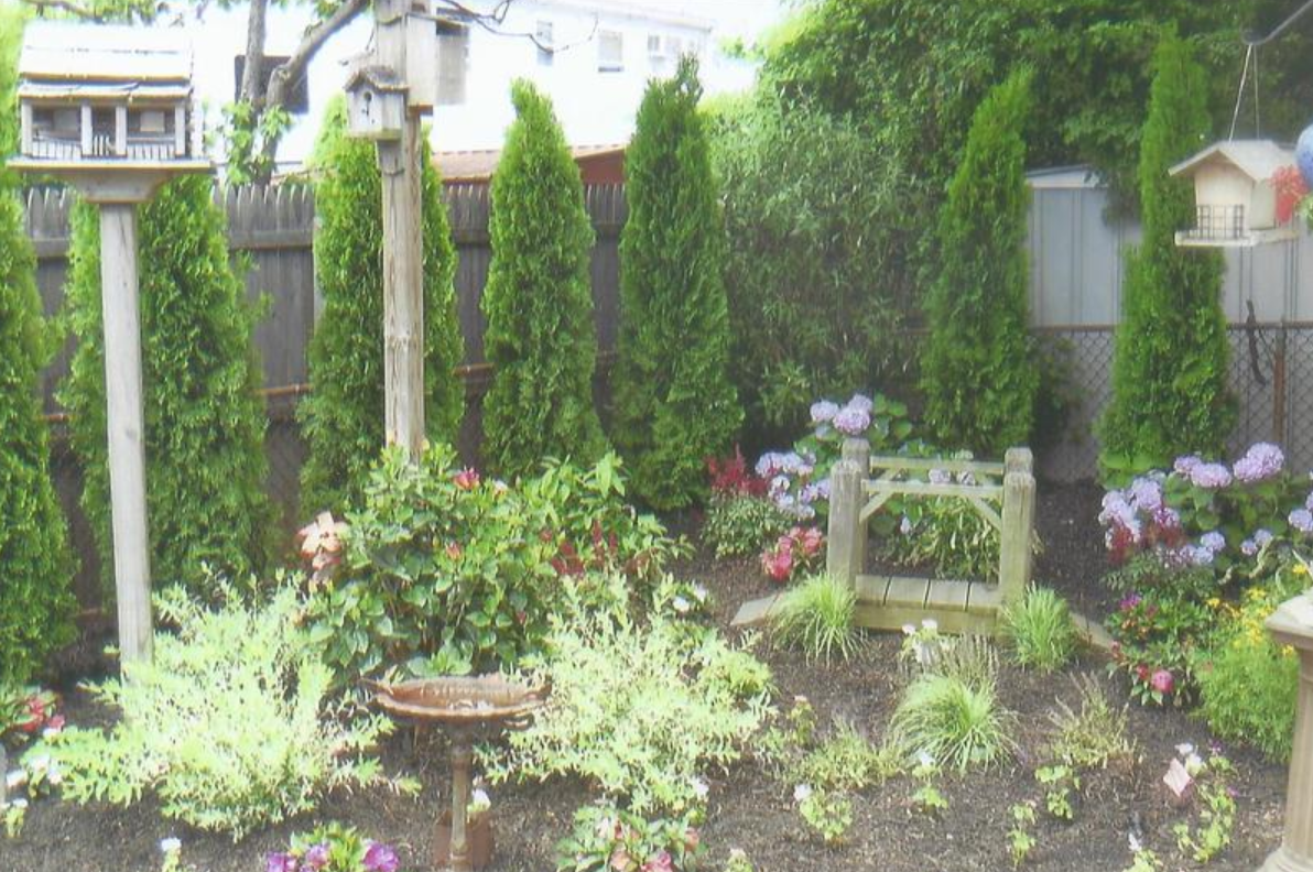A garden with evergreen trees, birdhouses, flowers, and a small wooden bridge.