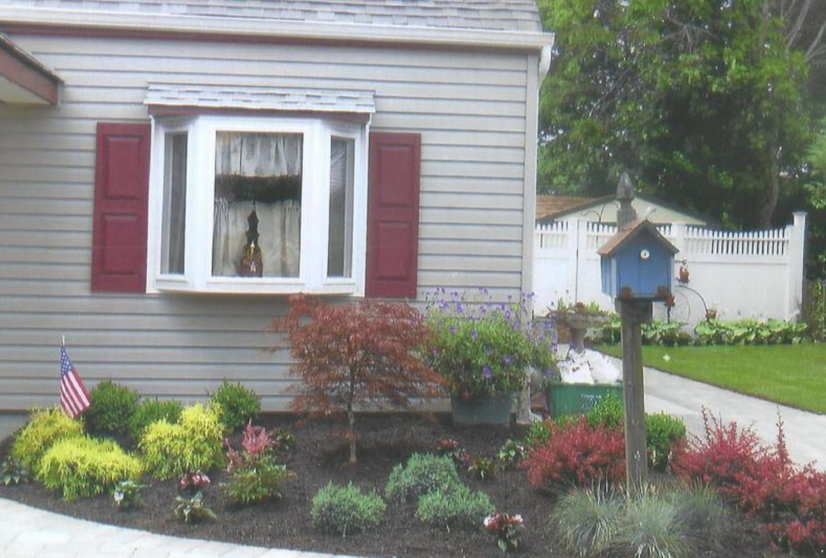 House exterior with a bay window, red shutters, a flower bed, and a blue birdhouse.