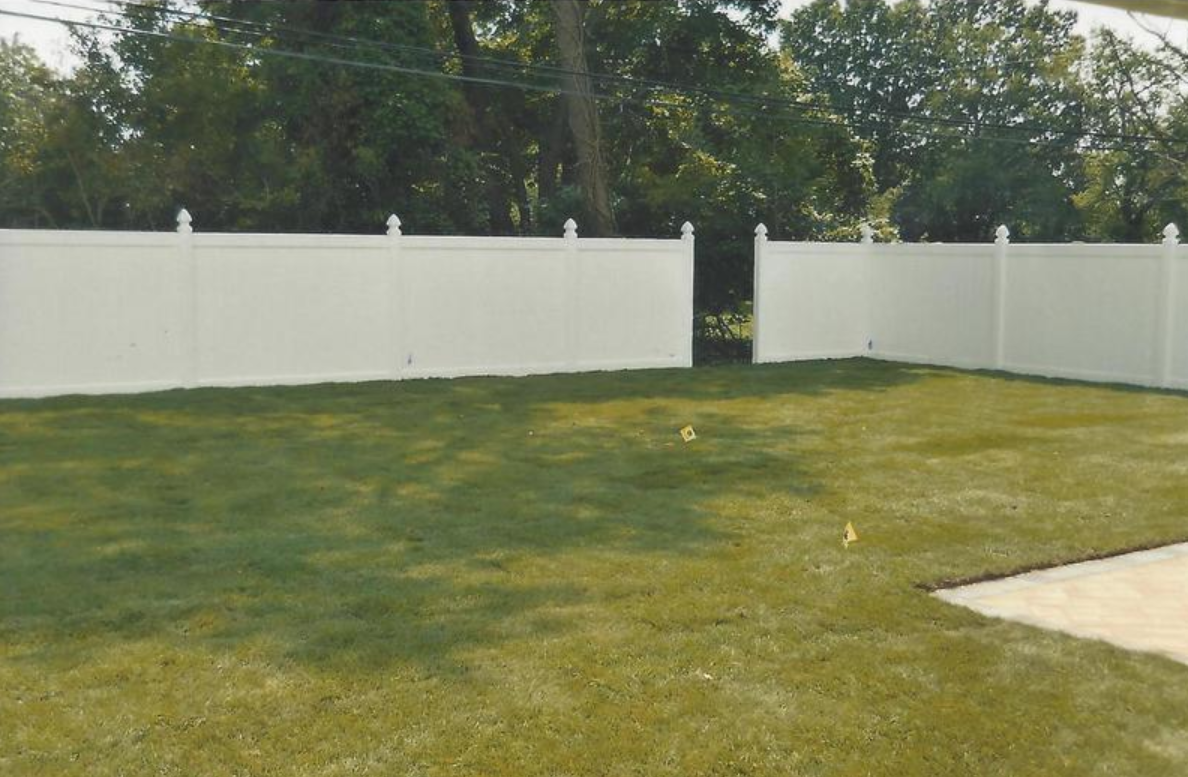 White vinyl fence surrounding a green grassy yard with an opening, trees in the background.