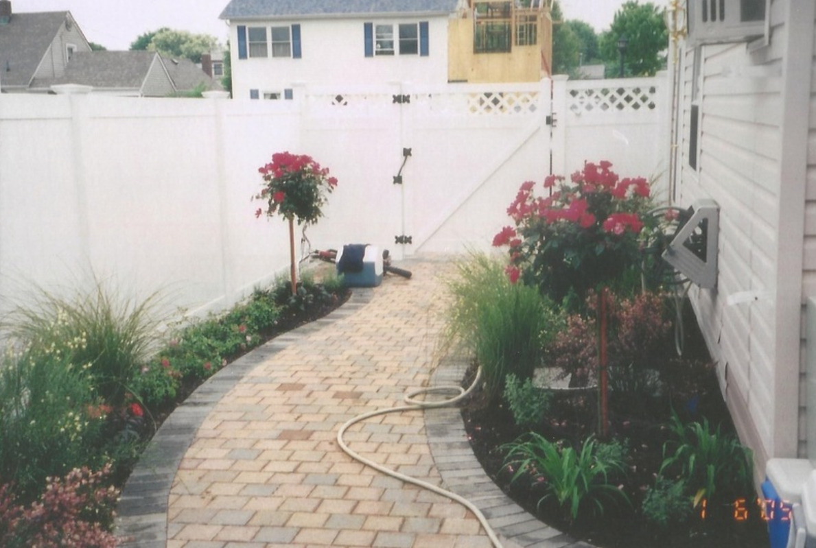 Brick pathway in a garden with flowers, leading to a white fence and gate.