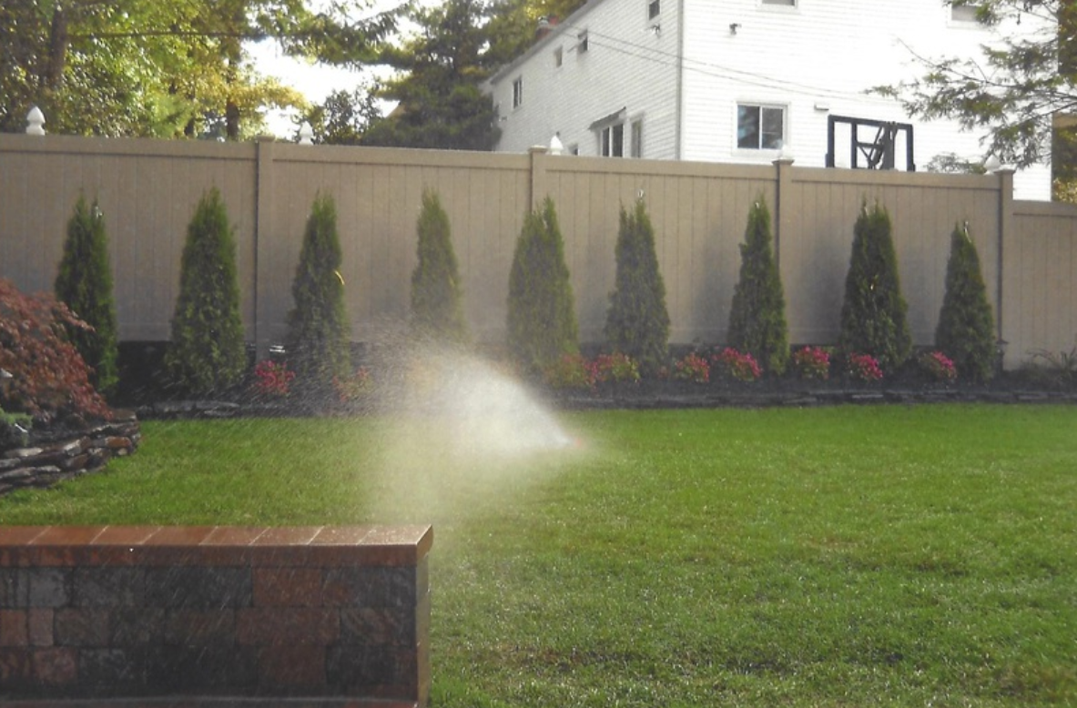 Lawn sprinkler spraying water in front of a tan fence, green grass, and evergreen trees.