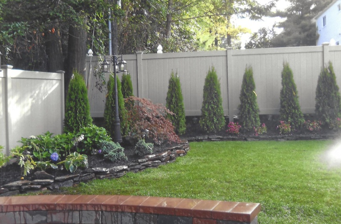 Backyard with green lawn, flower bed, trees, and white fence.