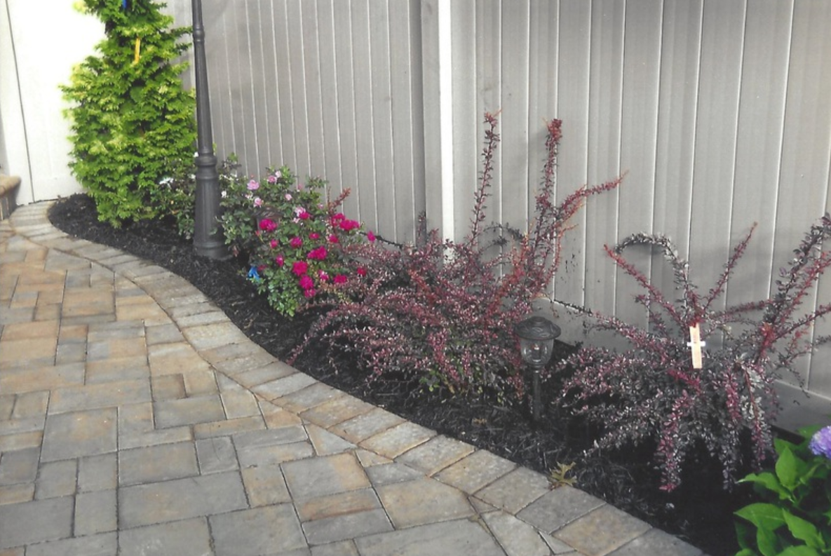 Brick pathway curves past a garden bed with dark red plants, flowers, and a green shrub against a gray fence.