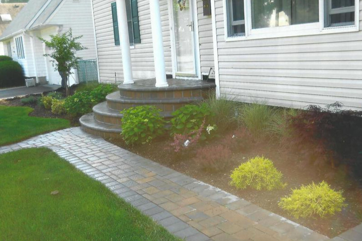 Brick pathway leading to a house with steps, flower beds, and green lawn.
