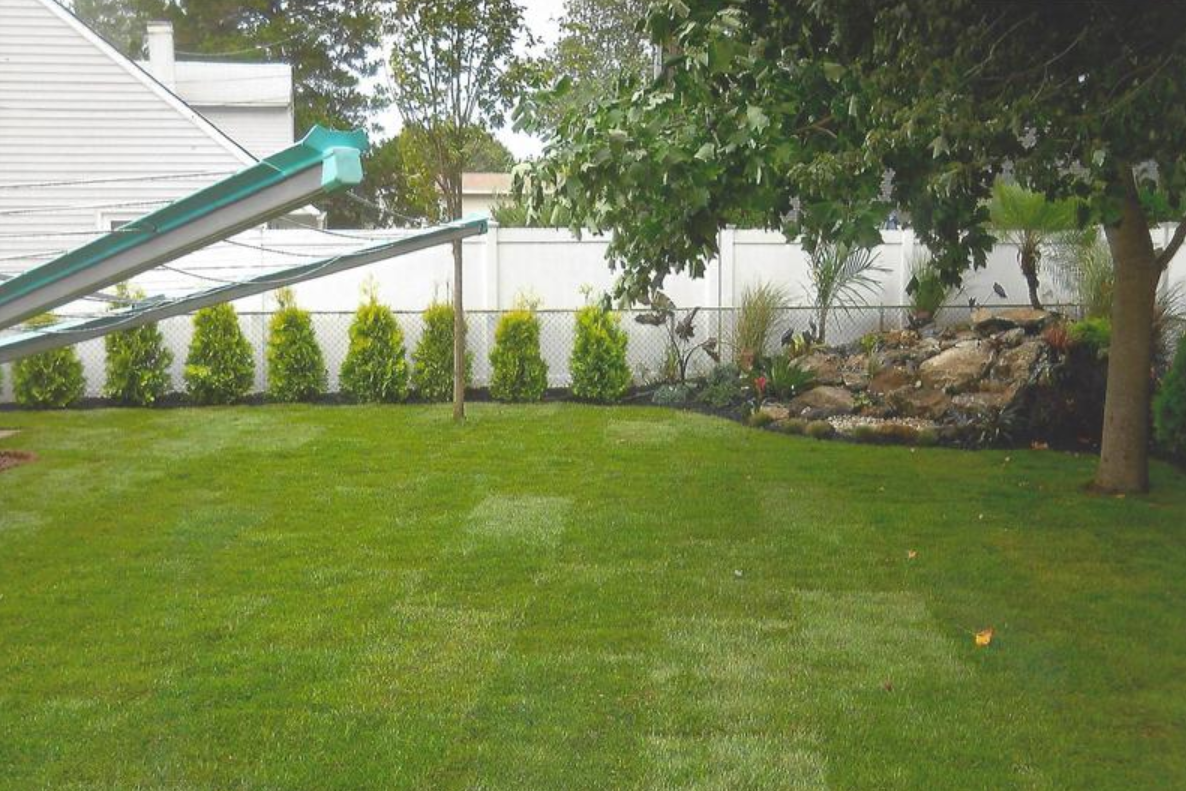 Green lawn in a backyard with a slide, trees, bushes, and a rock feature.