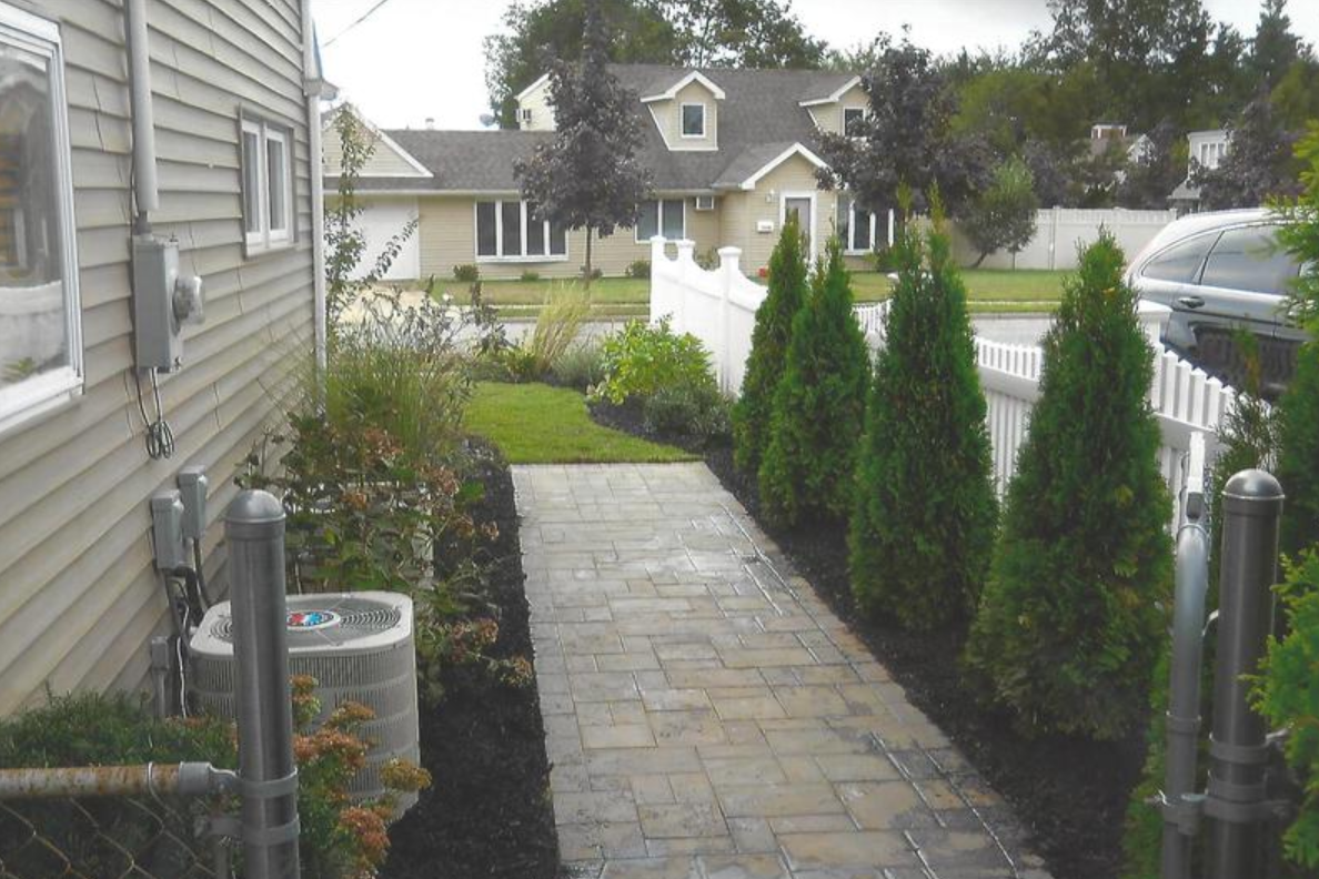 Stone path lined with green trees and white fence leading to a house with beige siding.