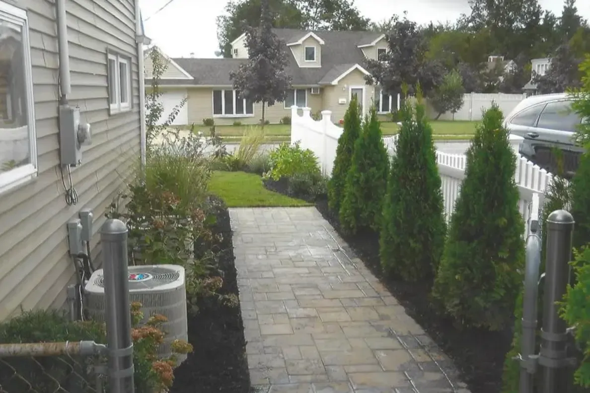 Brick walkway with evergreen trees, white fence, and beige houses.