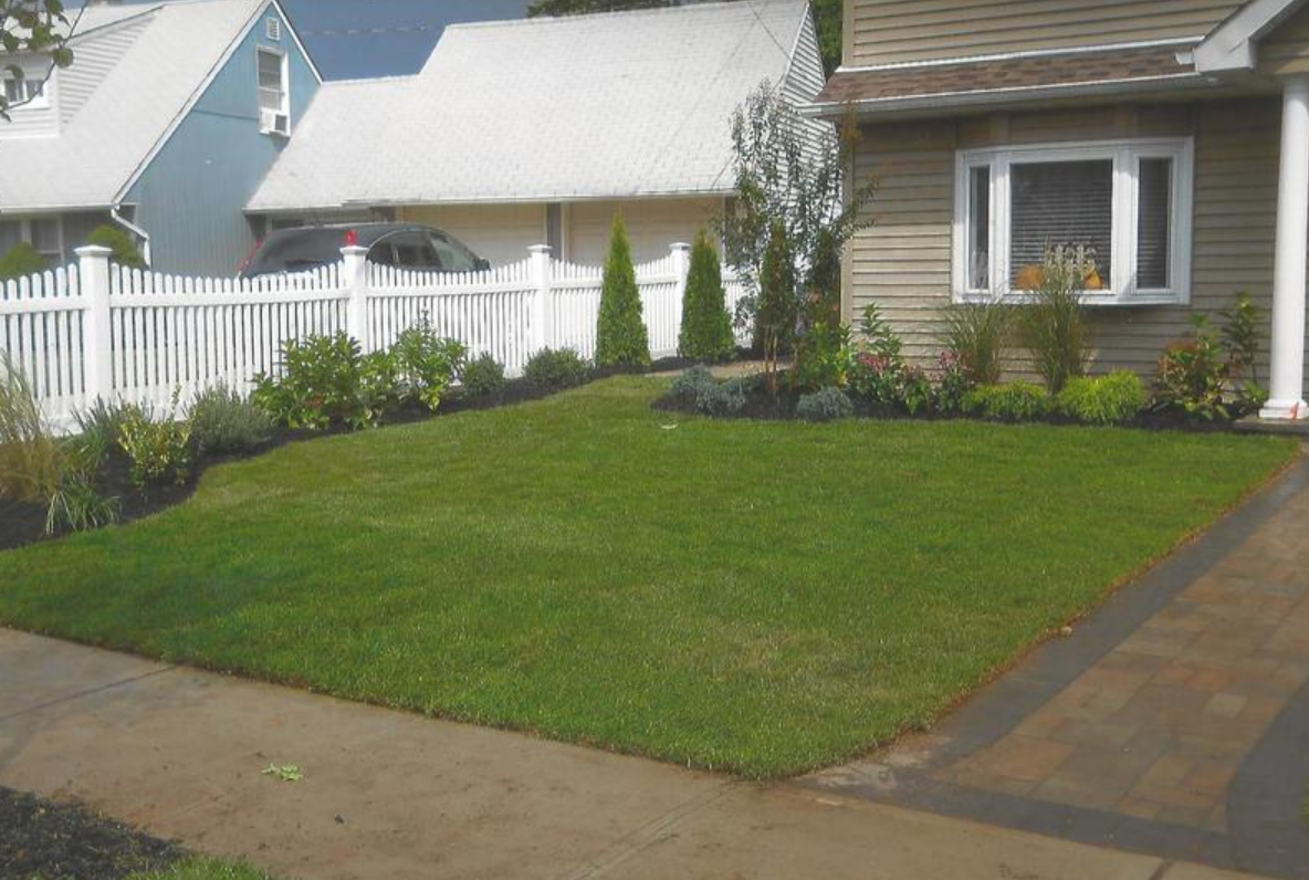 A well-manicured lawn in front of a beige house with landscaping, a white fence, and a blue house next door.