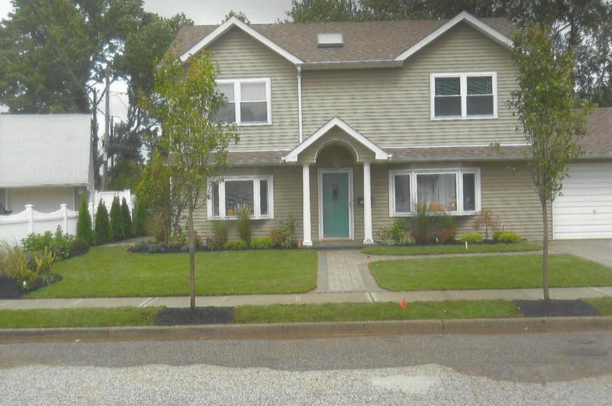 Two-story beige house with green door, green grass lawn, two trees, and gray driveway.