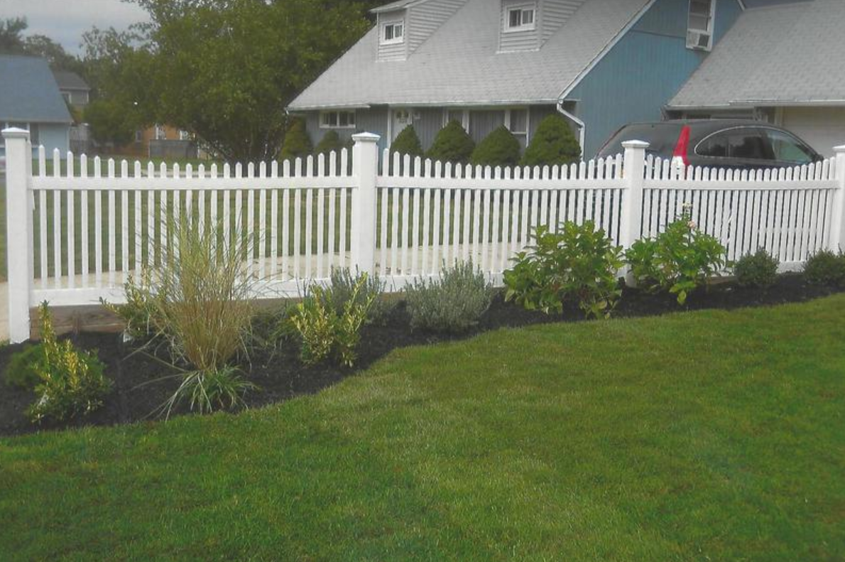 White picket fence in front of a house with a well-manicured lawn and landscaping.