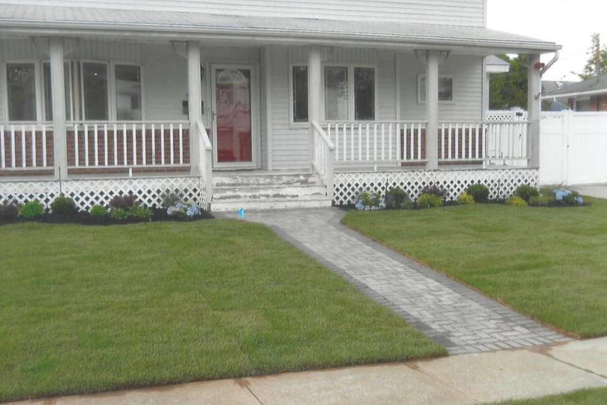 White house with a porch, red door, and brick pathway, surrounded by green grass and landscaping.