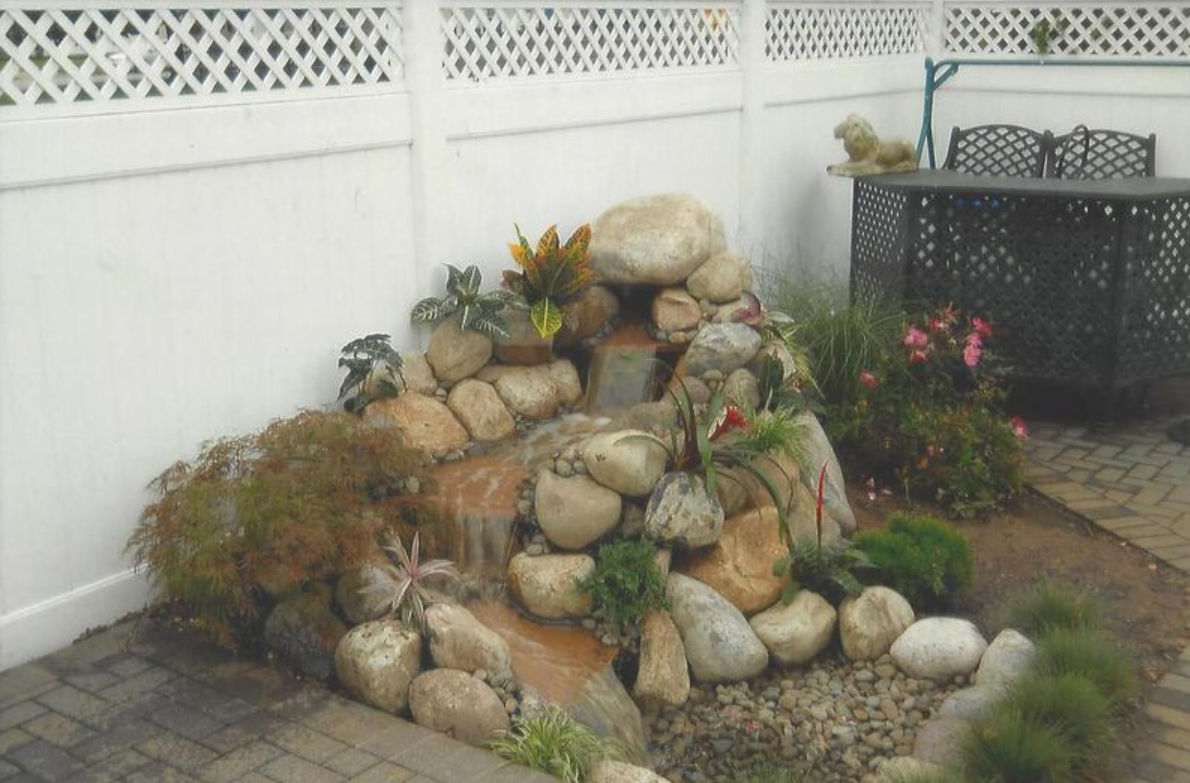 Waterfall feature with rocks and plants in a corner of a patio, with a white lattice fence behind it.