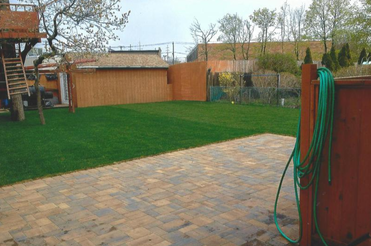 A backyard with green grass, a brown fence, wooden shed, and patio. A green hose hangs on the fence.