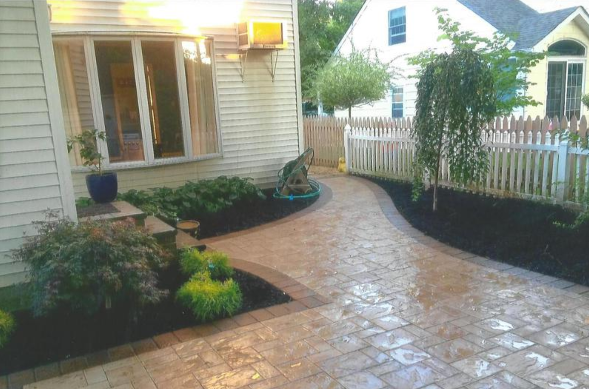 Brick pathway curves towards a white picket fence and house with landscaping.