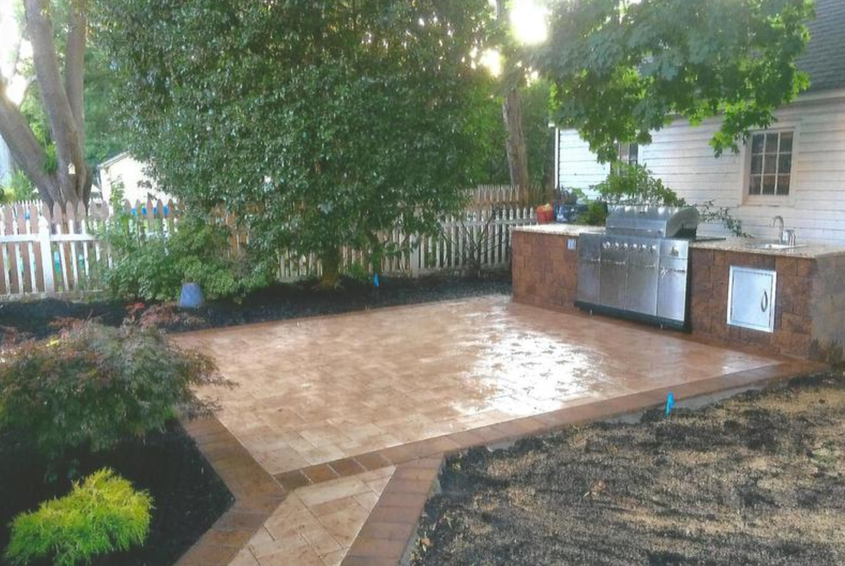 Brick patio with outdoor kitchen next to a white house.  Trees and fence in background.