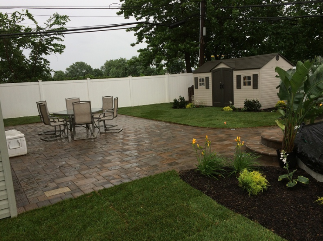 Backyard patio with table and chairs, a shed, and flower beds; white fence, green grass, and overcast sky.