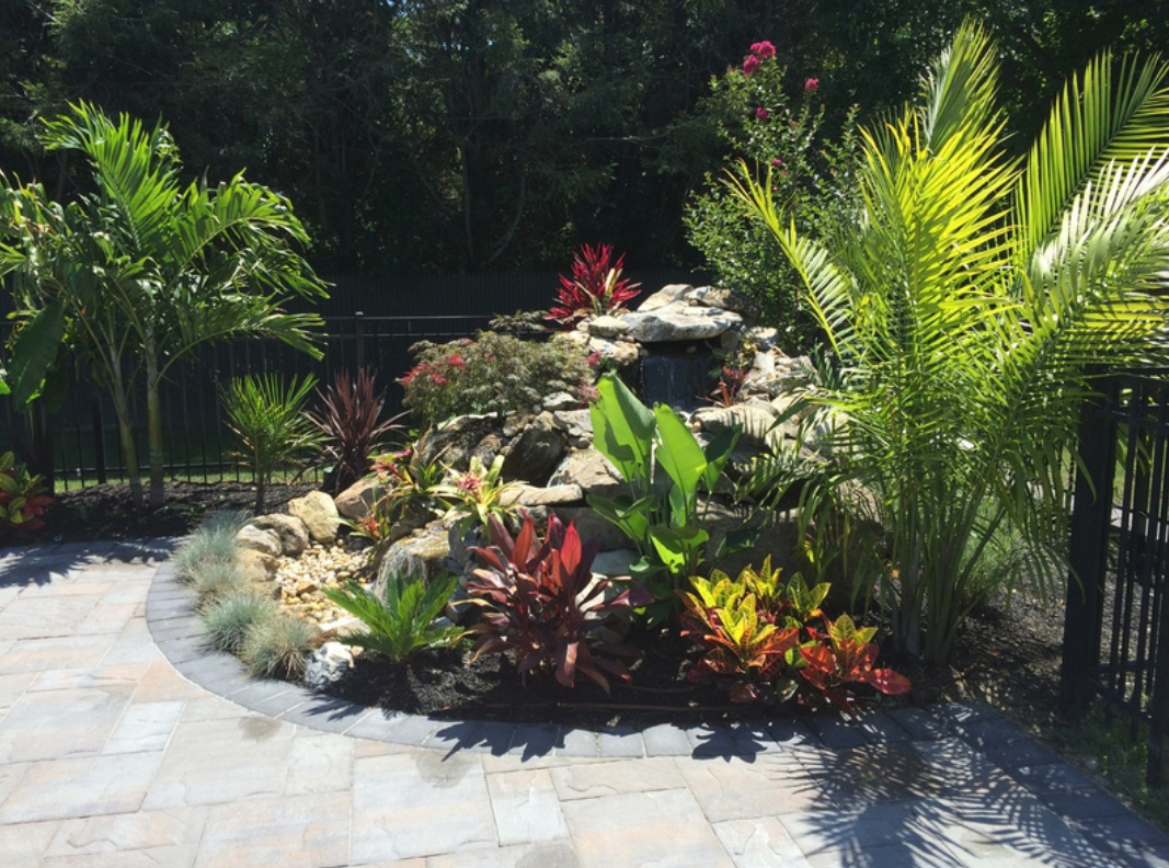 Lush tropical landscaping with a rock waterfall feature and colorful plants next to a paved patio.