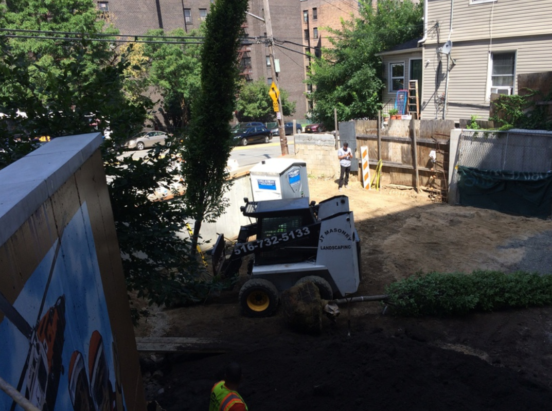 A Bobcat on dirt, with a porta-potty, and a person near a fenced area. Houses and a street in the background.