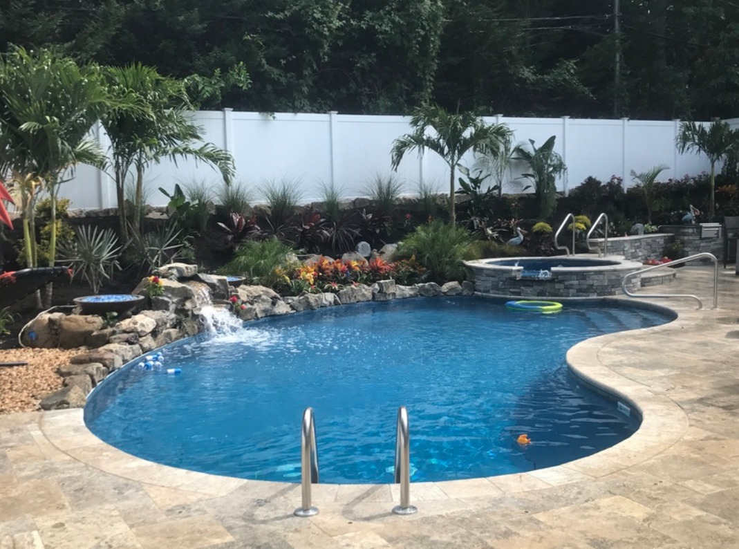 Swimming pool with a waterfall, surrounded by landscaping and a hot tub, against a white fence.