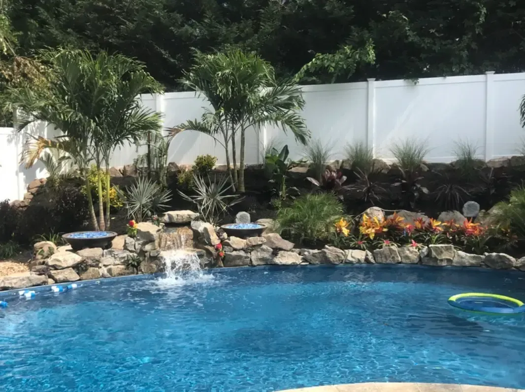 Pool with waterfall feature, landscaping, palm trees, and white fence in background.