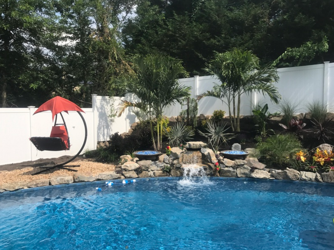 Swimming pool with waterfall feature, hanging chair, lush landscaping, and white fence.