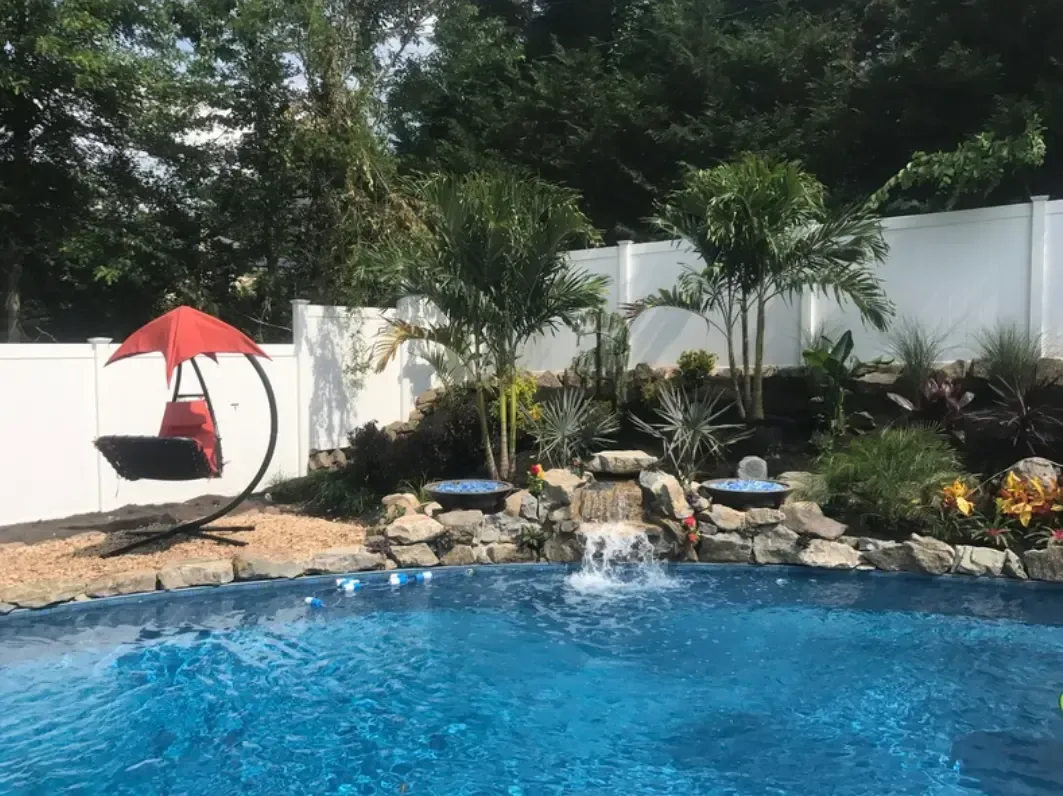 Pool with waterfall, landscaping, and a red swing chair; white fence and trees in the background.