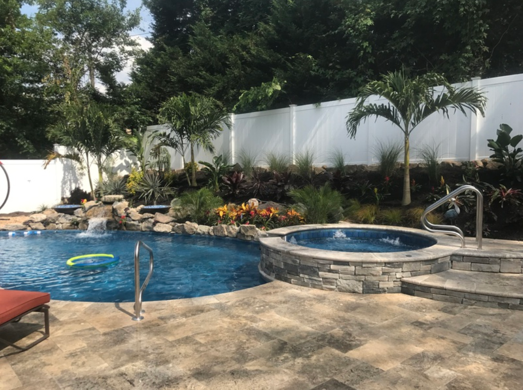 Backyard with a pool, jacuzzi, waterfall, and landscaping. A white fence and trees surround the area.