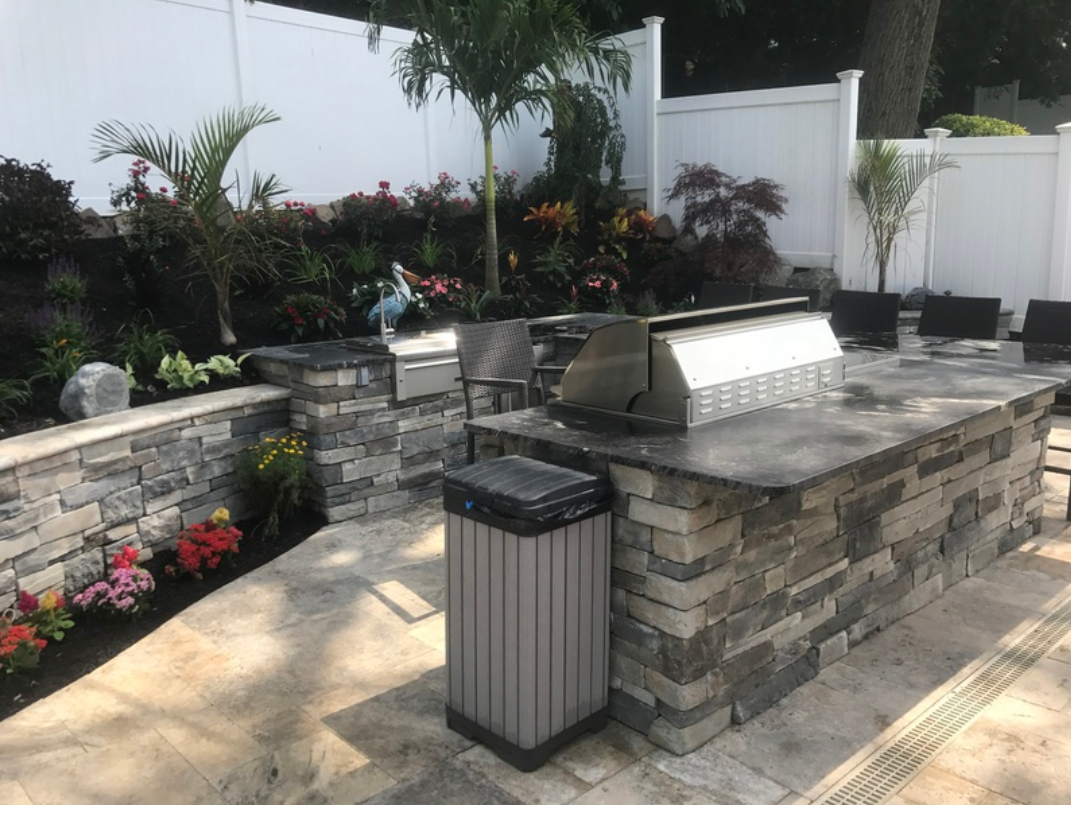 Outdoor kitchen with grill, stonework, and plants. Trash can in front. White fence in the background.