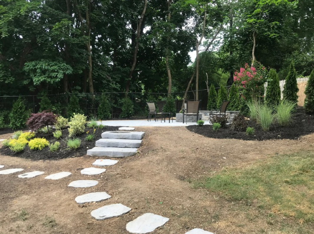 Stone path and steps lead up to a patio with seating, surrounded by plants and trees.