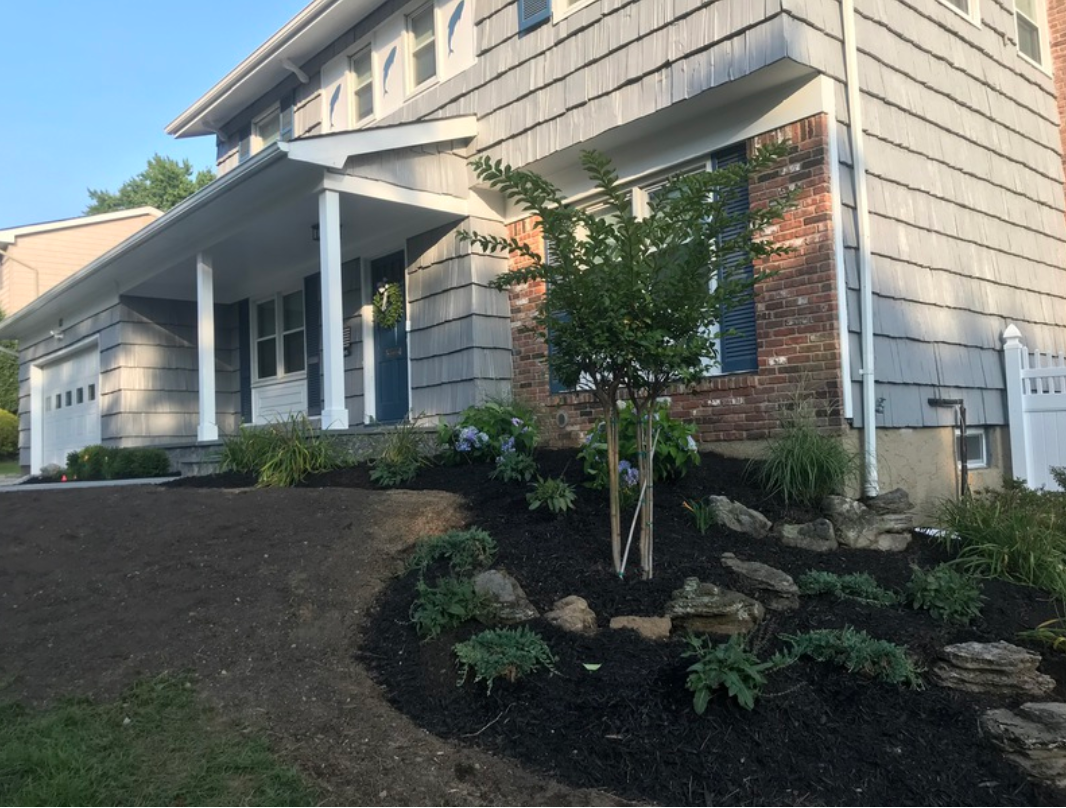 Gray house with dark mulch landscaping, featuring a young tree, rocks, and plants.