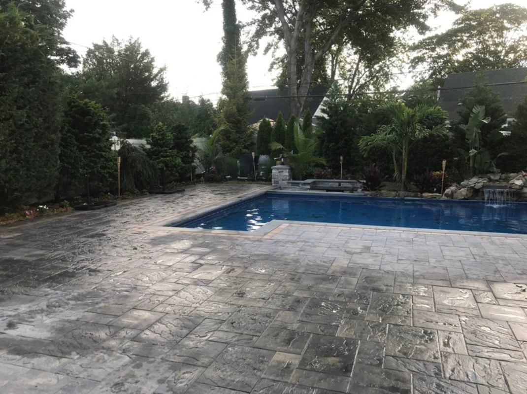 Backyard pool with paved patio, surrounded by green trees and shrubs. Overcast sky.