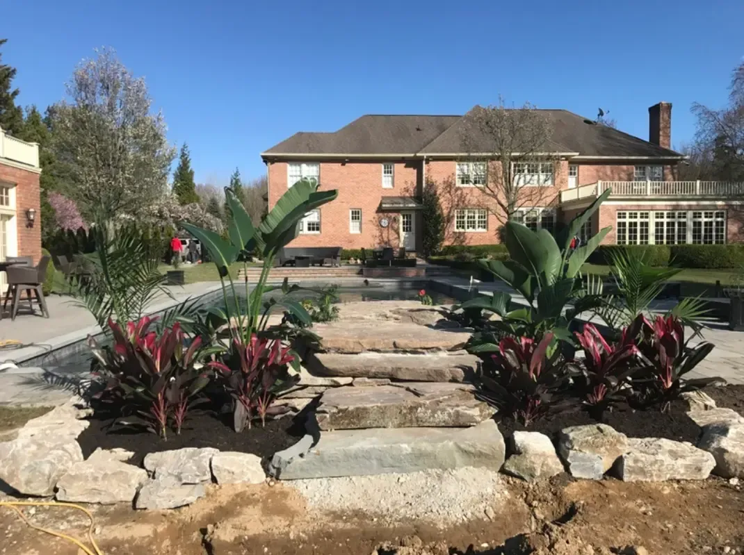 Stone steps and vibrant plants lead to a red-brick house under a blue sky.