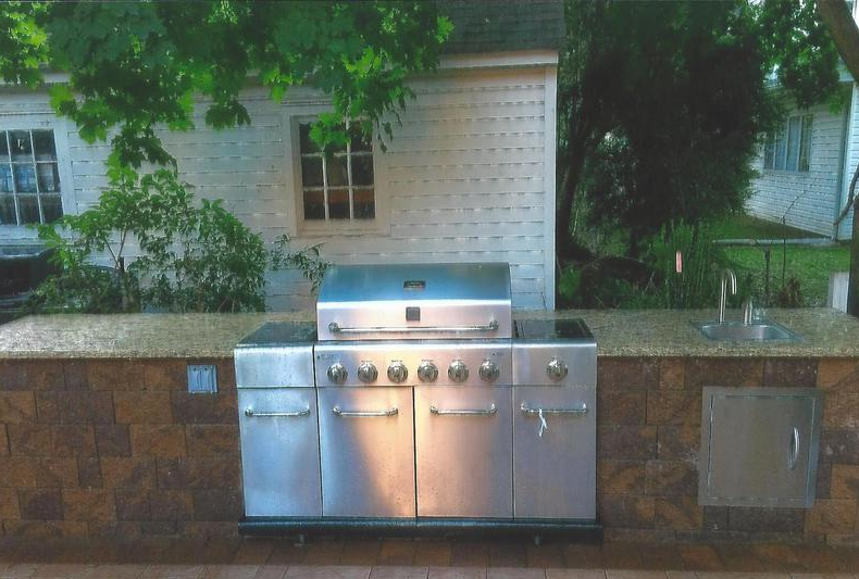 Outdoor kitchen with stainless steel grill, brown brick facade, and stone countertop.