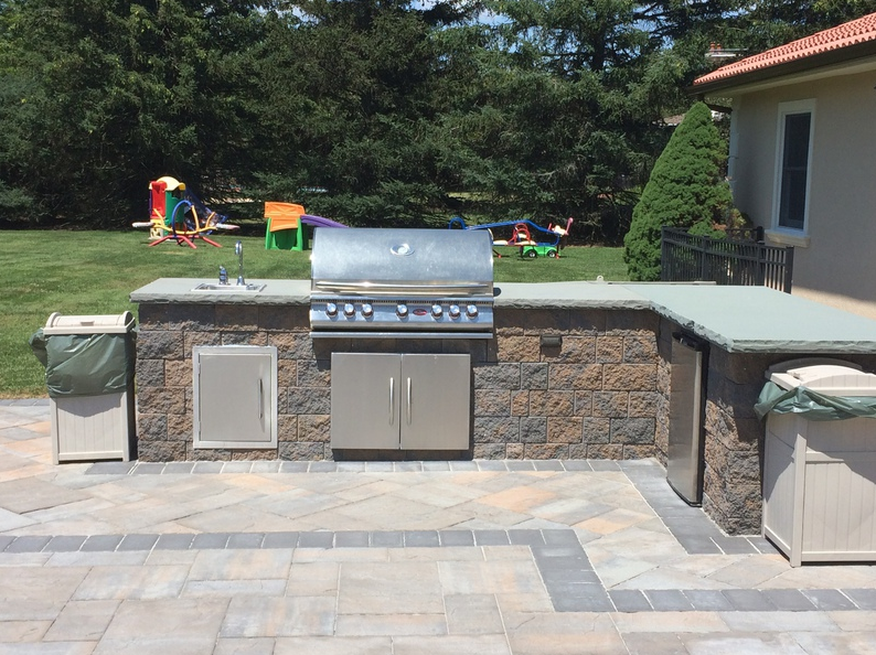 Outdoor kitchen with built-in grill, sink, and fridge.  Brown stone structure on a paved patio with lawn in the background.