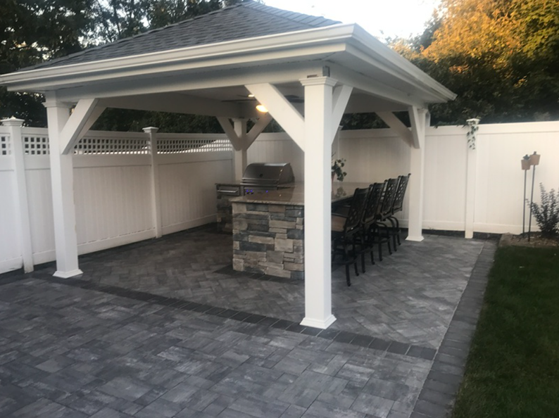 White gazebo with stone-faced grill and dining area, set on a brick patio, enclosed by a white fence.
