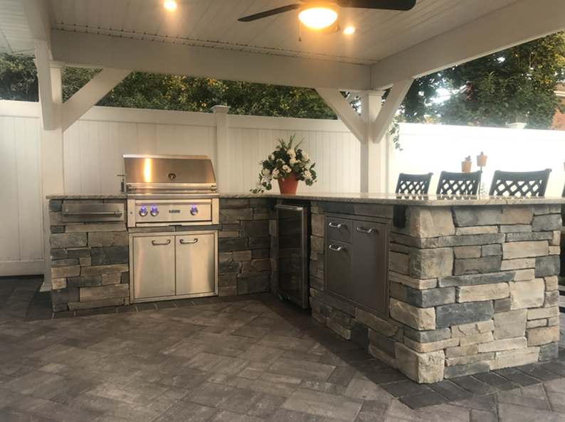 Outdoor kitchen with stone facade, stainless steel grill, bar, and seating under a white pergola.