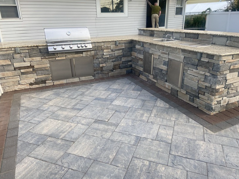Outdoor kitchen with stone facade, built-in grill and sink, on a paved patio.