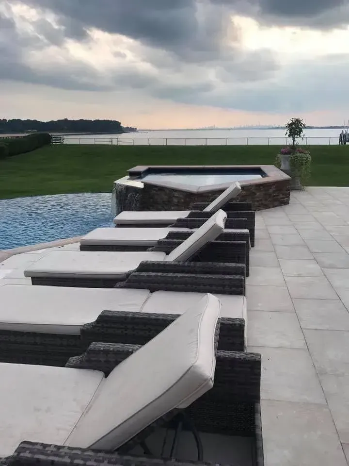Lounge chairs face a pool and hot tub overlooking a body of water under a cloudy sky.