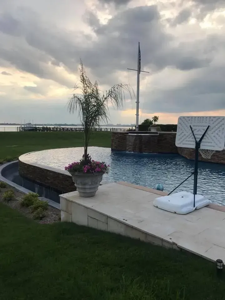 Poolside view with a basketball hoop, water feature, potted plant, and cloudy sky.