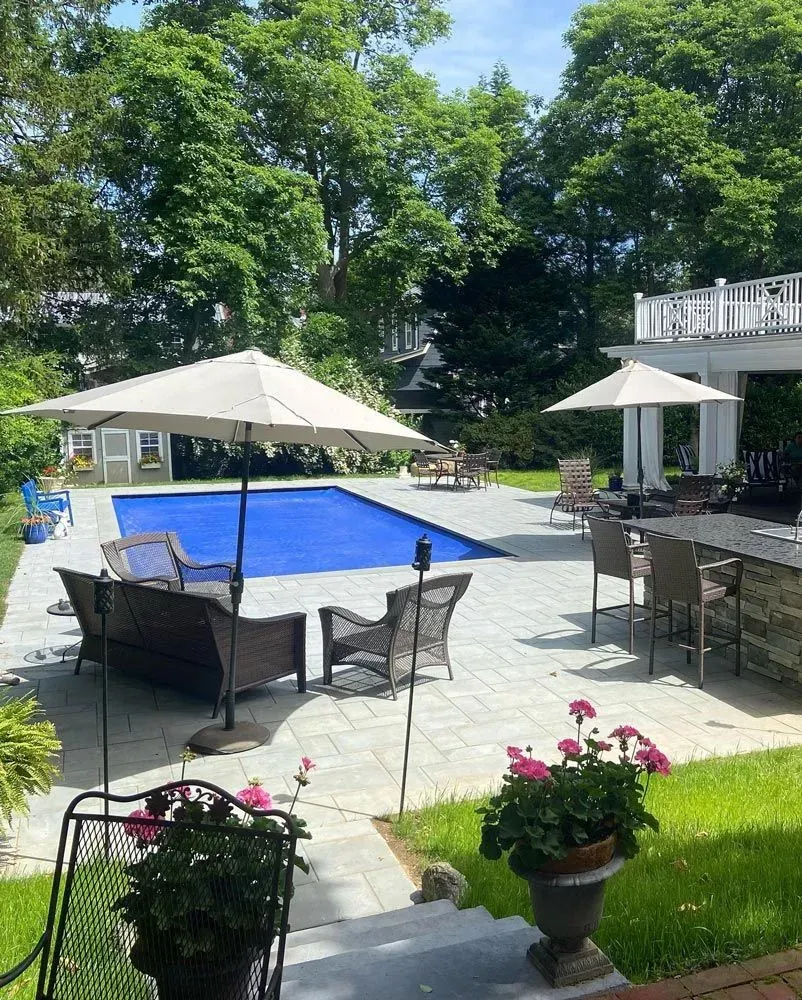 Backyard patio with pool, seating, and umbrellas on a sunny day.