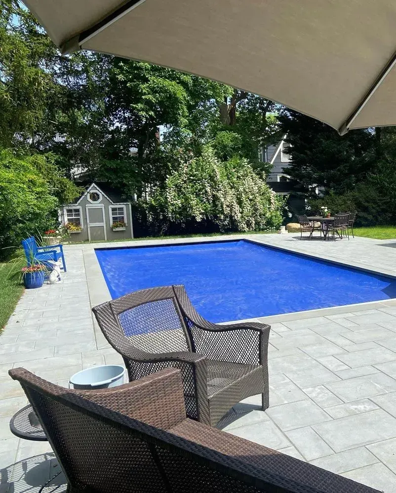 Blue-covered pool in a yard, with patio furniture and a small shed. Trees and umbrella provide shade.