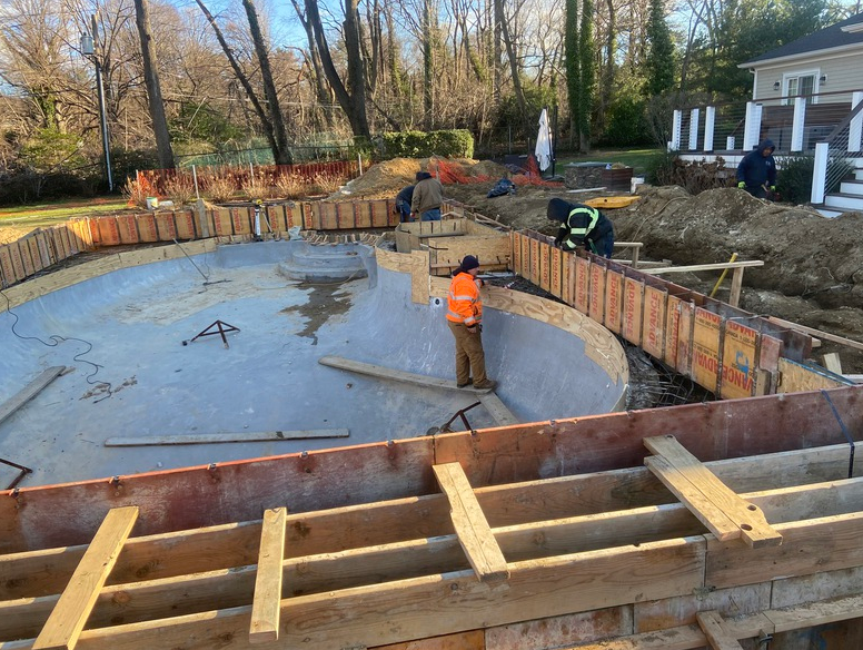 Pool construction site: Workers building formwork around concrete pool basin, with surrounding dirt and trees.