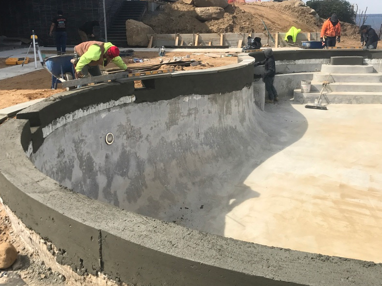 Construction workers building a curved pool. Concrete work in progress with workers applying cement. Outdoor setting.