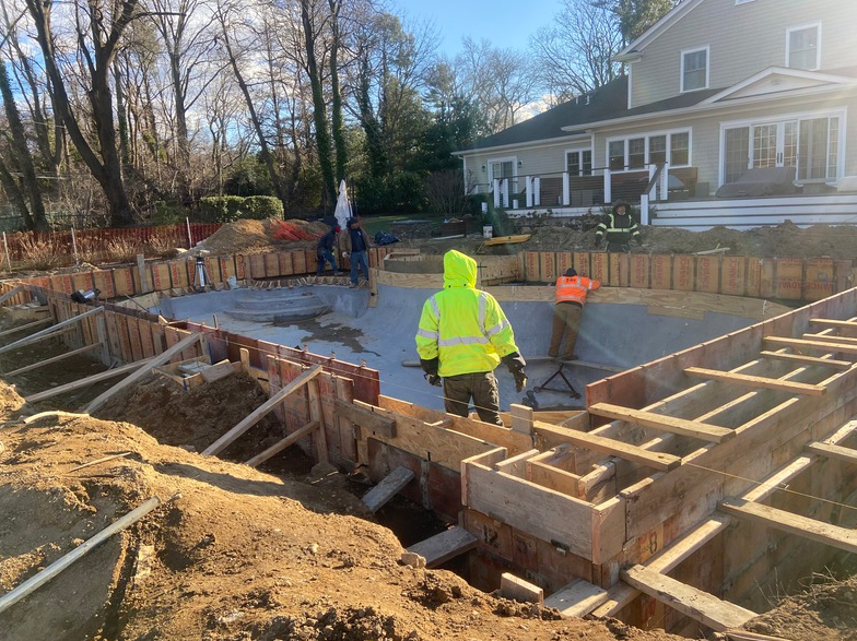 Construction site for a swimming pool. Workers in safety vests around wooden forms, a house in the background.