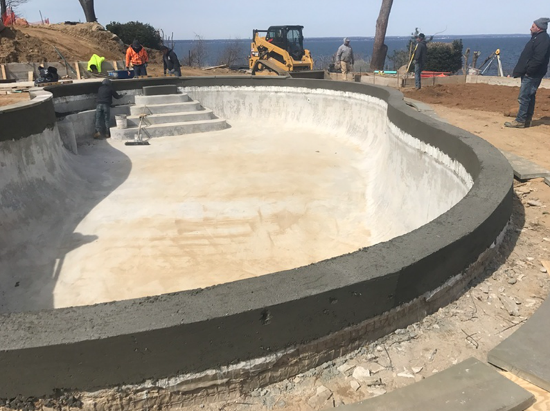 Construction of a concrete swimming pool. Workers, excavator, ocean backdrop.