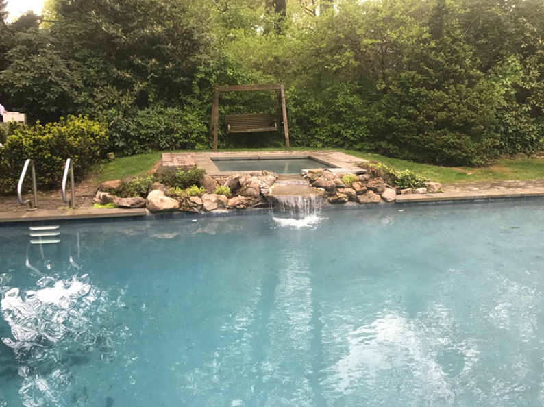A pool with a waterfall feature, jacuzzi, and swing, surrounded by greenery.