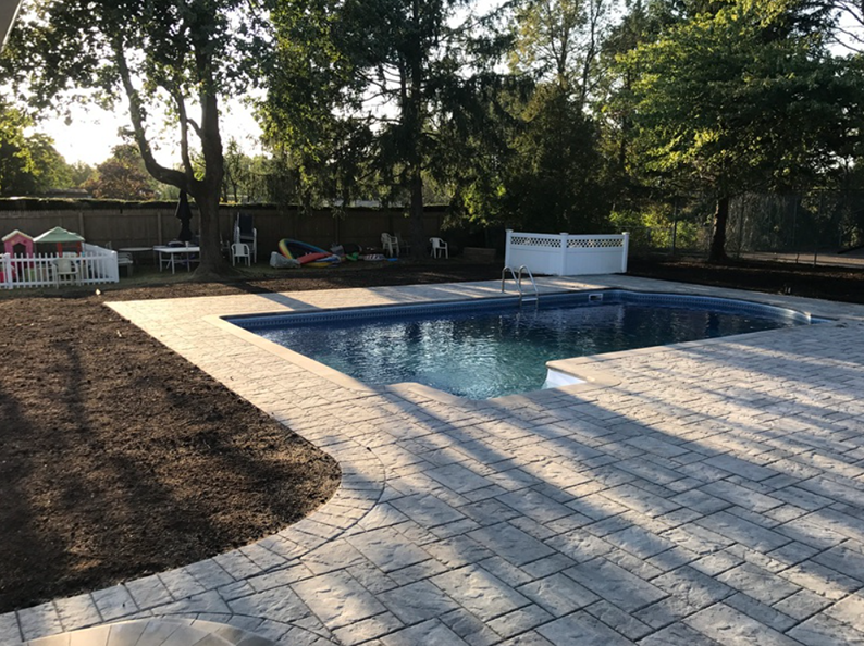 Backyard with a rectangular pool, brick patio, trees, and grassy area on a sunny day.