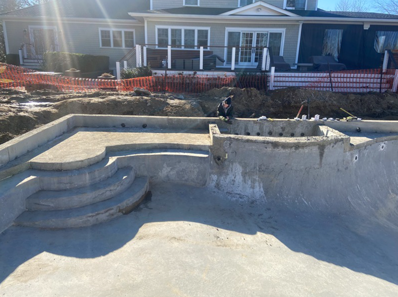 Concrete pool under construction with steps, surrounded by dirt and safety fencing. A person works nearby.